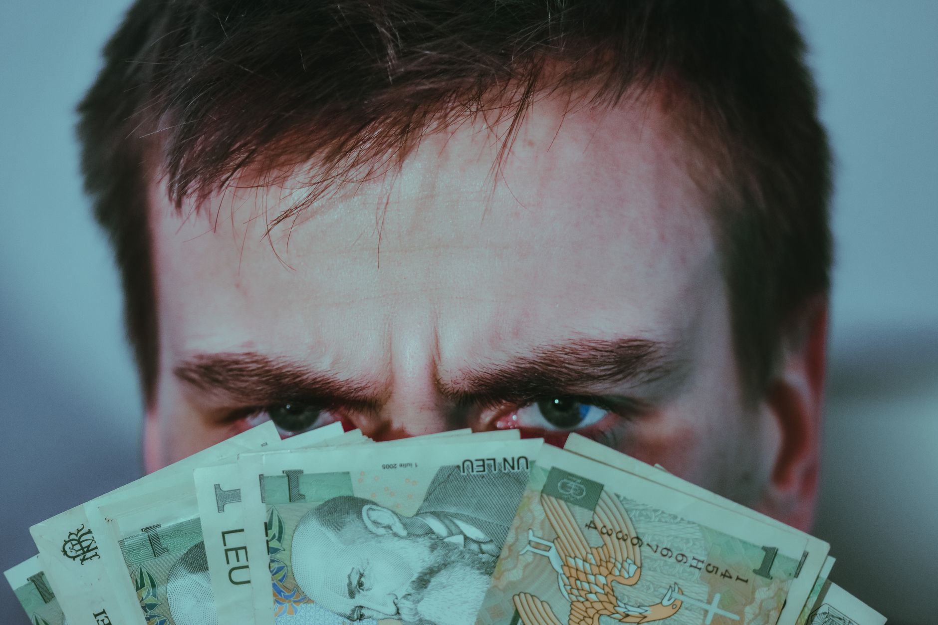 Crop unrecognizable frown man with dark hair covering face with stack of Romanian leu banknotes and looking at camera