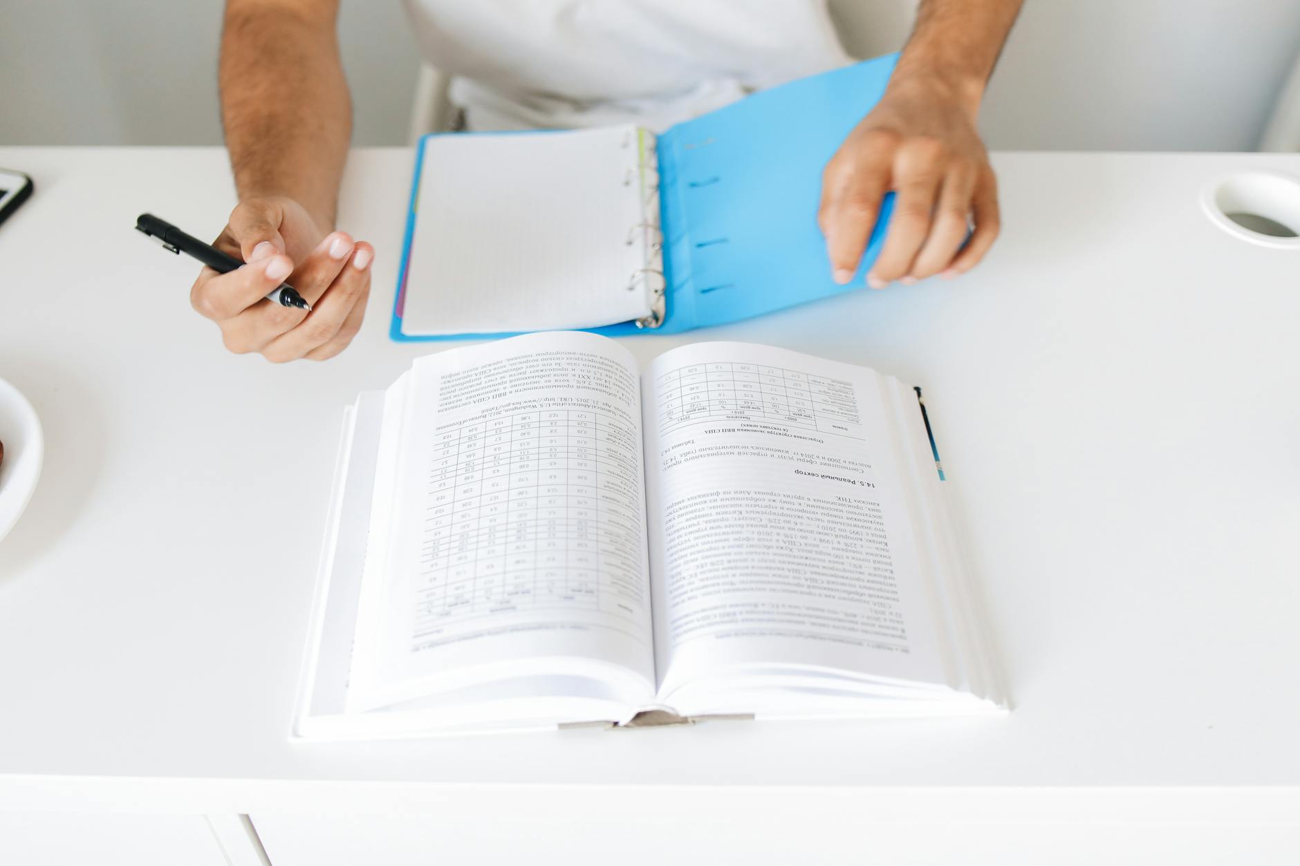 Close-up of a person taking notes from an open book and binder on desk.